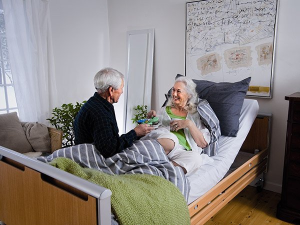 Seniorin liegt lächelnd im Pflegebett, Partner sitzt am Bett